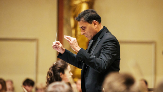 Vijay Upadhyaya stands in profile, his conducting wand raised and his head bowed as he consult the sheet music before him.